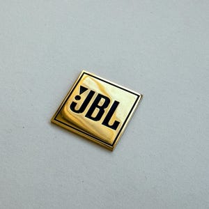 Peut inclure: Un embl&egrave;me carr&eacute; dor&eacute; avec le logo JBL en lettres noires. L'embl&egrave;me est l&eacute;g&egrave;rement r&eacute;fl&eacute;chissant et poss&egrave;de un bord noir. Le fond est de couleur gris clair.