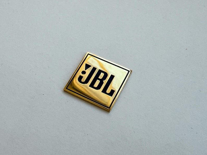 Peut inclure: Un embl&egrave;me carr&eacute; dor&eacute; avec le logo JBL en lettres noires. L'embl&egrave;me est l&eacute;g&egrave;rement r&eacute;fl&eacute;chissant et poss&egrave;de un bord noir. Le fond est de couleur gris clair.