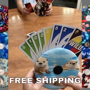 May include: A collection of round fabric card holders with button closures, each featuring unique patterns and designs. One holder displays otters in water, while others showcase floral, butterfly, and patriotic themes. The image also shows Uno cards.