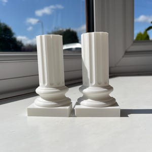 Pode incluir: Duas colunas coríntias brancas em miniatura com fustes canelados e bases detalhadas. As colunas estão sobre bases quadradas, mostrando um design arquitetônico clássico. A imagem é tirada com luz natural.