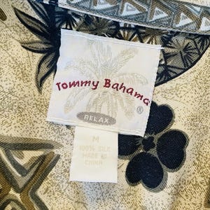 May include: Close-up of a Tommy Bahama shirt tag. The tag is white with the brand name in red script and a palm tree logo. The shirt fabric has a floral and geometric pattern in shades of tan, black, and green. The tag reads "RELAX" and "100% Silk".