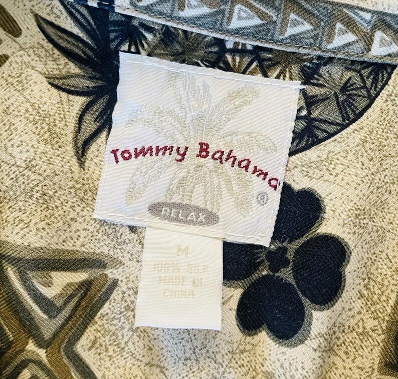 May include: Close-up of a Tommy Bahama shirt tag. The tag is white with the brand name in red script and a palm tree logo. The shirt fabric has a floral and geometric pattern in shades of tan, black, and green. The tag reads "RELAX" and "100% Silk".