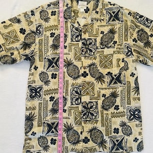 May include: Tan button-down shirt with a repeating pattern of pineapples, geometric shapes, and floral designs in shades of black, olive green, and tan. The shirt is approximately 30 inches long.