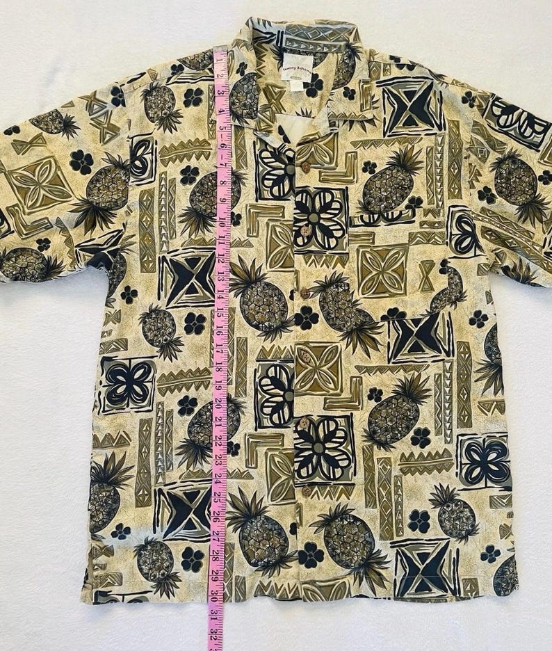 May include: Tan button-down shirt with a repeating pattern of pineapples, geometric shapes, and floral designs in shades of black, olive green, and tan. The shirt is approximately 30 inches long.