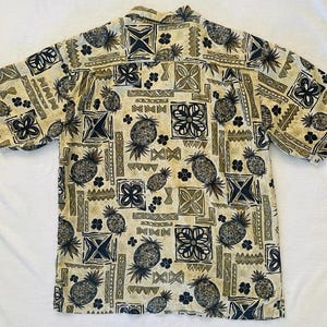 May include: Short-sleeved button-down shirt with a repeating pattern of pineapples, geometric shapes, and floral designs in shades of tan, olive green, and navy blue. The shirt has a classic collar and is made of a lightweight fabric.