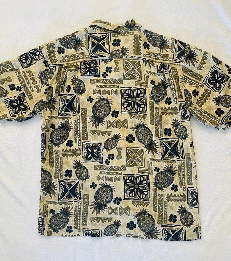 May include: Short-sleeved button-down shirt with a repeating pattern of pineapples, geometric shapes, and floral designs in shades of tan, olive green, and navy blue. The shirt has a classic collar and is made of a lightweight fabric.