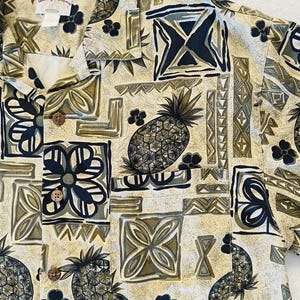 May include: A button-down shirt with a tropical pattern featuring pineapples, flowers, and geometric shapes in shades of beige, olive green, and navy blue. The shirt has a classic collar and wooden buttons. The brand name "Tommy Bahama" is visible.