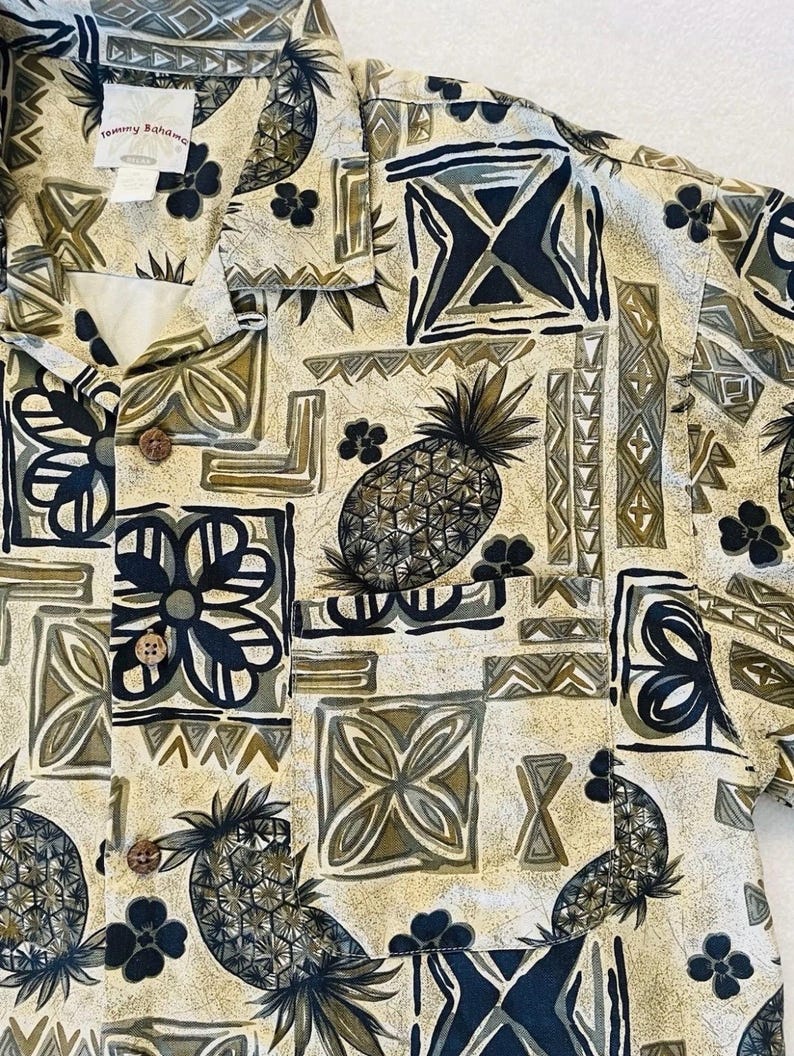 May include: A button-down shirt with a tropical pattern featuring pineapples, flowers, and geometric shapes in shades of beige, olive green, and navy blue. The shirt has a classic collar and wooden buttons. The brand name "Tommy Bahama" is visible.