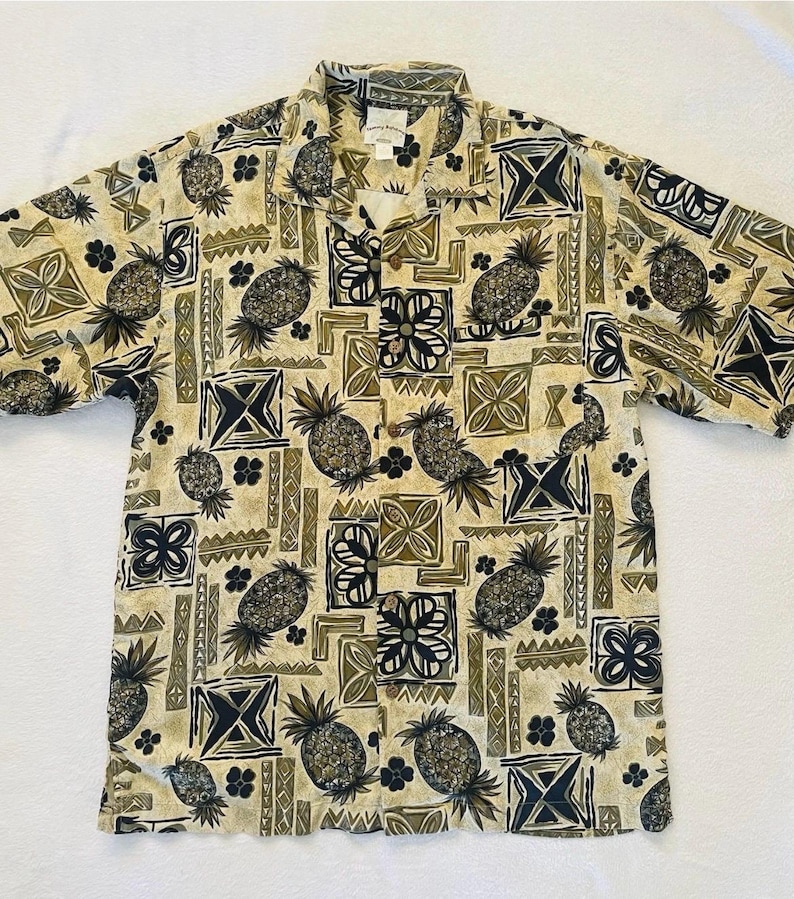 May include: Short-sleeved button-down shirt with a repeating pattern of pineapples, geometric shapes, and floral designs in shades of beige, black, and olive green. The shirt has a collared neckline and a relaxed fit, suitable for casual wear.