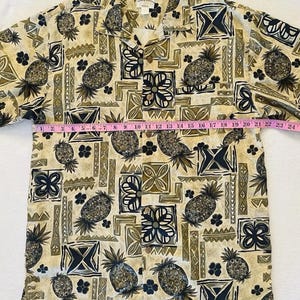 May include: Tan button-down shirt with a repeating pattern of pineapples, flowers, and geometric shapes in shades of brown, black, and green. The shirt has a collared neckline and short sleeves. The shirt measures approximately 24 inches across.