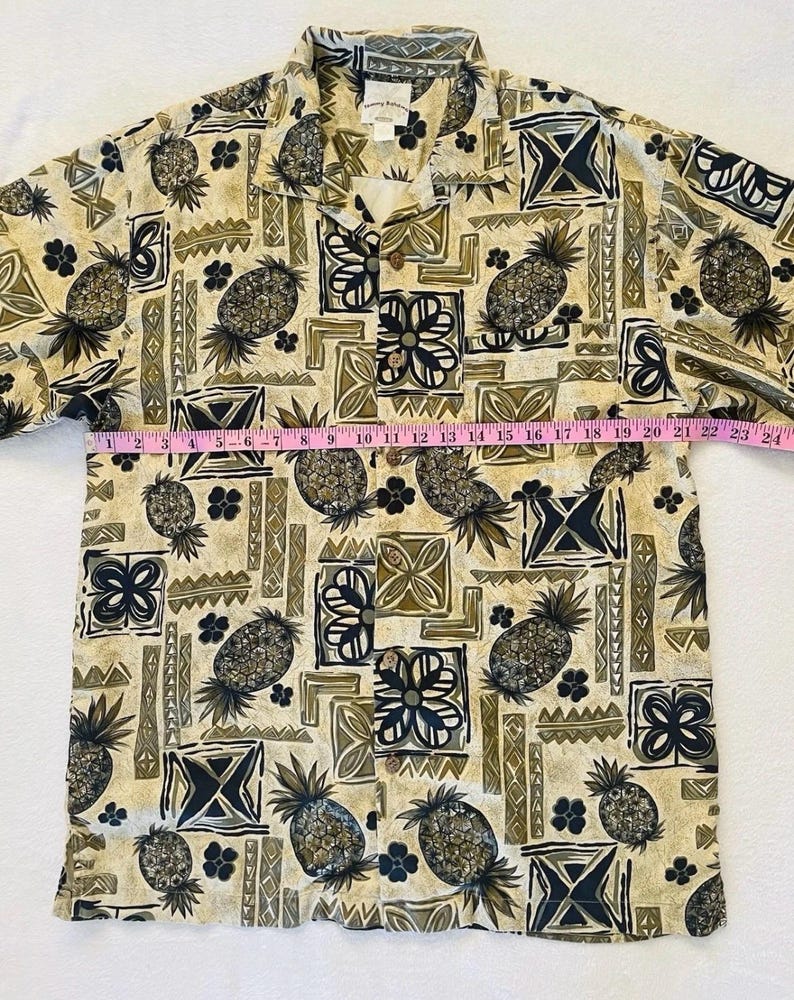 May include: Tan button-down shirt with a repeating pattern of pineapples, flowers, and geometric shapes in shades of brown, black, and green. The shirt has a collared neckline and short sleeves. The shirt measures approximately 24 inches across.