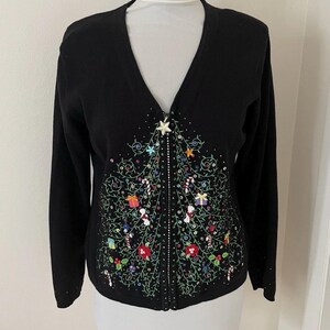Puede incluir: Cárdigan negro con cuello en V y diseño de árbol de Navidad. El árbol está adornado con lentejuelas, abalorios y detalles bordados de colores, incluyendo regalos, bastones de caramelo y estrellas. El cárdigan tiene cierre de cremallera y mangas largas.