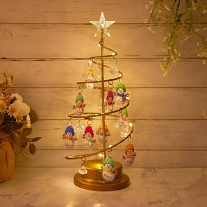May include: A gold-coloured, spiral Christmas tree with a star on top, decorated with snowman ornaments. The tree is lit with small lights and stands on a round base. The background is a light-coloured wooden wall.