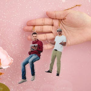 Könnte beinhalten: Zwei transparente Acryl-Anhänger mit Fotos von Männern. Ein Mann trägt ein rotes Hemd und hält ein Buch. Der andere Mann trägt ein weißes Hemd und eine Khakihose. Beide Anhänger haben eine goldene Schnur.