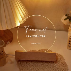 May include: A glowing, circular LED night light with the words "Fear not, I am with you" and the reference Isaiah 41:10. The light sits on a rectangular wooden base, creating a warm, inviting ambiance.
