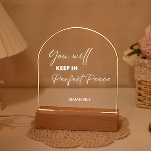 May include: A lighted acrylic sign with the words "You will keep in Perfect Peace" and the Bible verse Isaiah 26:3. The sign is mounted on a wooden base and sits on a decorative doily.