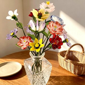 May include: A colorful glass flower bouquet in a clear, square-shaped vase. The flowers are various colors, including red, yellow, white, and purple. A small wicker basket and a plate are also visible.