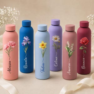 Op de afbeelding: Zes cilindervormige metalen waterflessen in diverse pastel- en felle kleuren, elk voorzien van een unieke bloemenillustratie en een gepersonaliseerde naam in scriptlettertype: Alessia, Renata, Talia, Celine, Amara en Bianca.