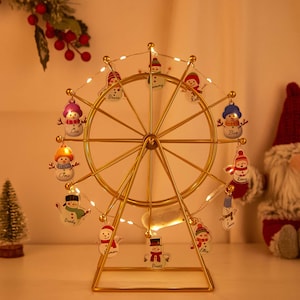 Peut inclure: Décoration de Noël en forme de grande roue dorée avec de petits ornements de bonhommes de neige. La roue est illuminée par de minuscules lumières blanches. Chaque bonhomme de neige porte un nom. Un petit sapin et un Père Noël sont en arrière-plan.