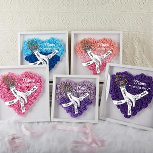 May include: Five framed heart-shaped floral arrangements in various colors: blue, pink, peach, purple, and lavender. Each features a hand holding a bouquet and the word "Mom" with names. The frames are white, and the background is a soft, textured fabric.