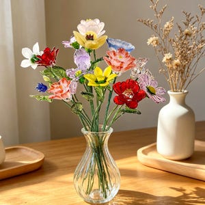 May include: A glass vase filled with colorful glass flowers. The flowers include red, yellow, pink, and white blooms with green stems. The vase is clear and sits on a wooden surface.