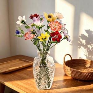 May include: A clear glass vase with a geometric pattern holds a colorful bouquet of glass flowers. The flowers include red, yellow, pink, and purple blooms with green stems. The vase sits on a wooden table, with a woven basket and wooden tray nearby.