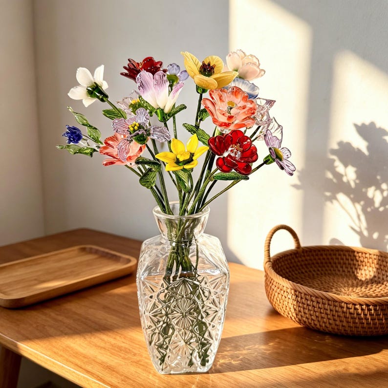 May include: A clear glass vase with a geometric pattern holds a colorful bouquet of glass flowers. The flowers include red, yellow, pink, and purple blooms with green stems. The vase sits on a wooden table, with a woven basket and wooden tray nearby.