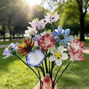 May include: A bouquet of colorful glass flowers in various shapes and sizes. The flowers include shades of pink, blue, white, orange, and purple. The stems are dark green, and the background is a blurred green lawn.