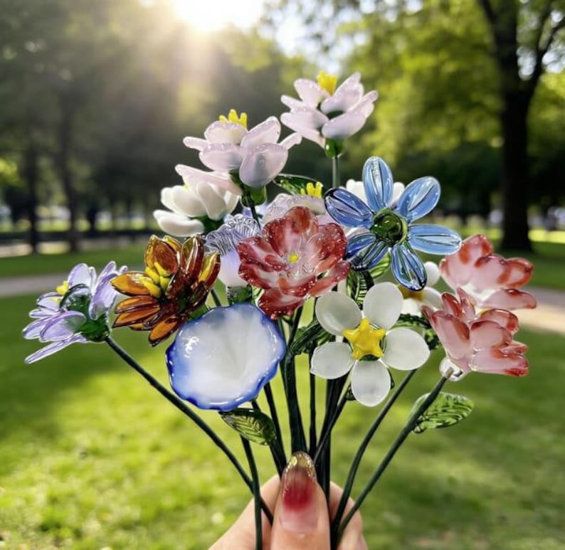 May include: A bouquet of colorful glass flowers in various shapes and sizes. The flowers include shades of pink, blue, white, orange, and purple. The stems are dark green, and the background is a blurred green lawn.