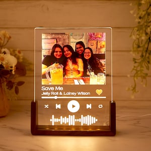 May include: A personalized acrylic night light with a photo of four people. The light has a wooden base and displays the song title "Save Me" by Jelly Roll & Lainey Wilson, along with a music player interface.