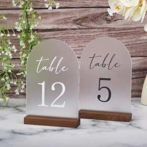 Può includere: Due cartelli numerati per tavolo in acrilico smerigliato con basi in legno. Il cartello a sinistra dice "tavolo 12" in testo bianco. Il cartello a destra dice "tavolo 5" in testo nero.