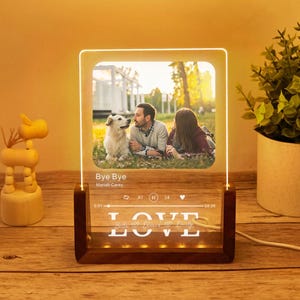 Może przedstawiać: Spersonalizowana lampka nocna LED ze zdjęciem pary i psa. Światło ma drewnianą podstawę z podświetlonym słowem "LOVE". Wyświetlana jest piosenka "Bye Bye" Mariah Carey.