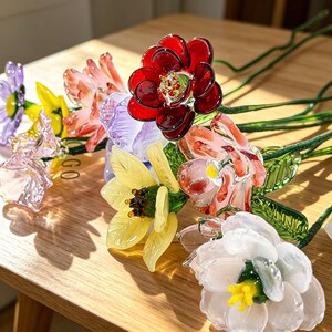 May include: A collection of colorful glass flowers in various hues, including red, yellow, purple, and white. The flowers have detailed petals and green stems, arranged on a wooden surface. The glass flowers are a decorative item.