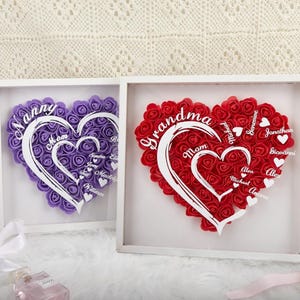 May include: Two framed heart-shaped floral arrangements. One is purple with the words "Nanny" and "Mom" and names. The other is red with the words "Grandma" and "Mom" and names. Both are in white frames.