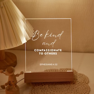 May include: A clear acrylic LED night light with the words "Be kind and compassionate to others" in white script. The light sits on a wooden base and includes the Bible verse Ephesians 4:32.