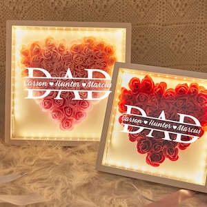 May include: Two illuminated heart-shaped floral arrangements. One features pink roses, the other red roses. The word "DAD" is displayed in white, with names below. The frames are white, with warm white LED lights.