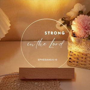 May include: A decorative LED lamp with a wooden base and a circular, illuminated design. The text "STRONG in the Lord" is displayed, along with "Ephesians 6:10". A basket of flowers is in the background.