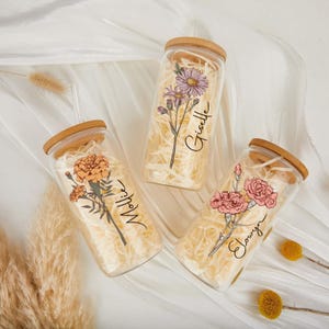 May include: Three glass tumblers with wooden lids, each featuring a floral design and a name. The flowers are in shades of orange, pink, and purple. The tumblers are filled with shredded paper.