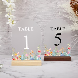 Puede incluir: Dos letreros arqueados con números de mesa. Los letreros tienen un fondo gris con la palabra "TABLE" en la parte superior y un número en el centro. La parte inferior de cada letrero está decorada con un diseño floral de acuarela. Un letrero está sobre una base de madera clara y el otro sobre una base de madera oscura.