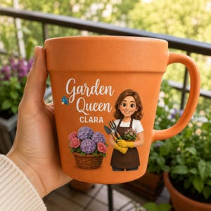 May include: A terracotta-colored ceramic mug shaped like a flower pot. The front features the text "Garden Queen" above the name "CLARA," a small blue bird, a basket of purple and pink hydrangeas, and an illustration of a person holding garden tools.