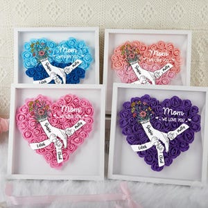 Peut inclure: Quatre arrangements floraux en forme de cœur encadrés, dans des tons de bleu, rose et violet. Chaque cœur présente un bouquet, le mot "Mom" et le texte "We Love You". Des rubans nominatifs personnalisés sont également inclus.