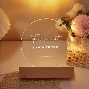 Op de afbeelding: Decoratieve LED-lamp met een houten voet. De heldere acryl cirkel toont de tekst "Fear not, I AM WITH YOU" en "ISAIAH 41:10". Op de achtergrond staat een bloemstuk. Geschikt voor interieurdecoratie.