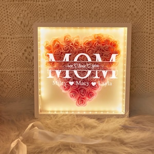 May include: White light-up frame with a heart-shaped arrangement of pink and peach artificial roses. The text "MOM" is displayed in a white font with the words "we love you" and the names "Miley, Macy, Layla" below.