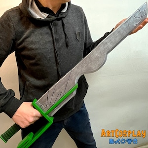 Puede incluir: Una espada grande de color plateado con un mango verde. La hoja tiene un patrón decorativo y un borde curvo. La espada está sostenida por una persona que lleva una sudadera gris y pantalones vaqueros azules.