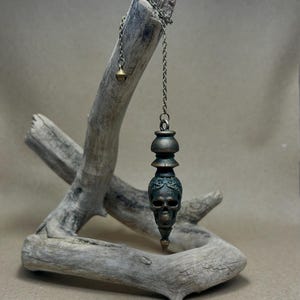 May include: A bronze-toned metal pendulum featuring a detailed skull carving at the base, topped with a decorative tiered cap. The pendulum hangs from a thin silver-colored metal chain that includes a small spherical bead at the end.