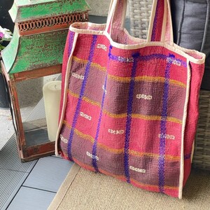 K&ouml;nnte beinhalten: Eine gro&szlig;e, rote und violette Tragetasche mit Karomuster und wei&szlig;em Besatz. Die Tasche ist aus gestepptem Stoff und hat einen breiten, flachen Boden.