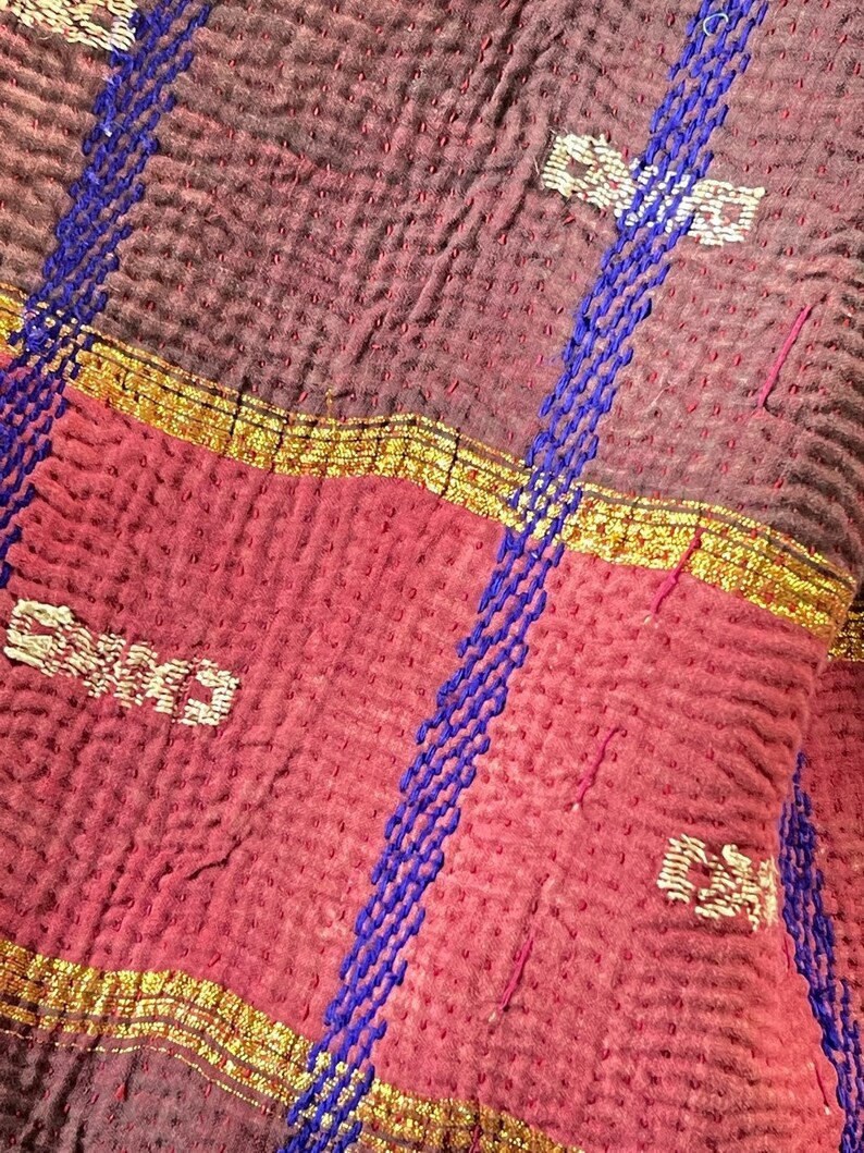 K&ouml;nnte beinhalten: Nahaufnahme einer roten und braunen Kantha-Steppdecke mit blauen und goldenen Stickereien. Die Steppdecke besteht aus mehreren Stofflagen, die zusammengen&auml;ht werden, um einen gesteppten Effekt zu erzielen. Die Stickerei ist aufwendig und detailliert, und die Farben sind leuchtend und kr&auml;ftig.