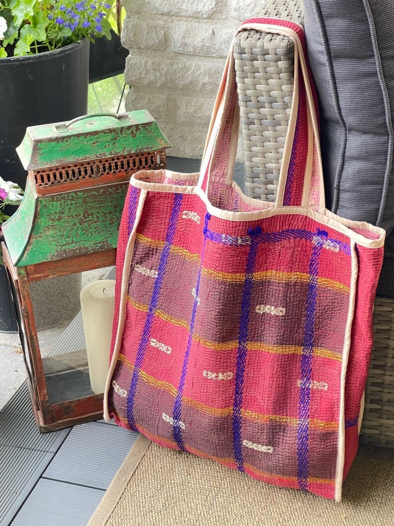 K&ouml;nnte beinhalten: Eine gro&szlig;e, rote und violette Tragetasche mit Karomuster und wei&szlig;em Besatz. Die Tasche ist aus gestepptem Stoff und hat einen breiten, flachen Boden.