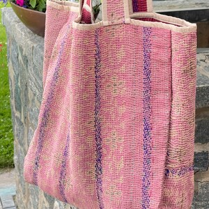K&ouml;nnte beinhalten: Eine rosafarbene und blaue gesteppte Tragetasche mit einem gestreiften Muster. Die Tasche hat eine gro&szlig;e Tasche an der Seite und zwei lange Henkel.
