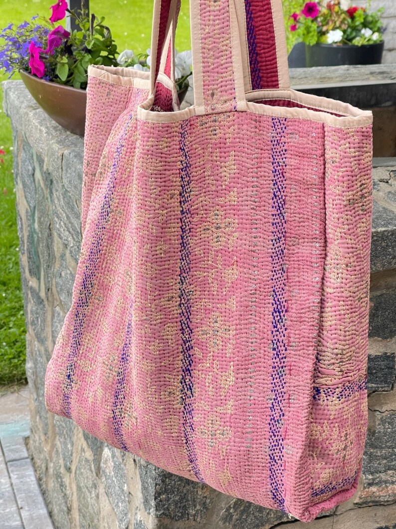 K&ouml;nnte beinhalten: Eine rosafarbene und blaue gesteppte Tragetasche mit einem gestreiften Muster. Die Tasche hat eine gro&szlig;e Tasche an der Seite und zwei lange Henkel.